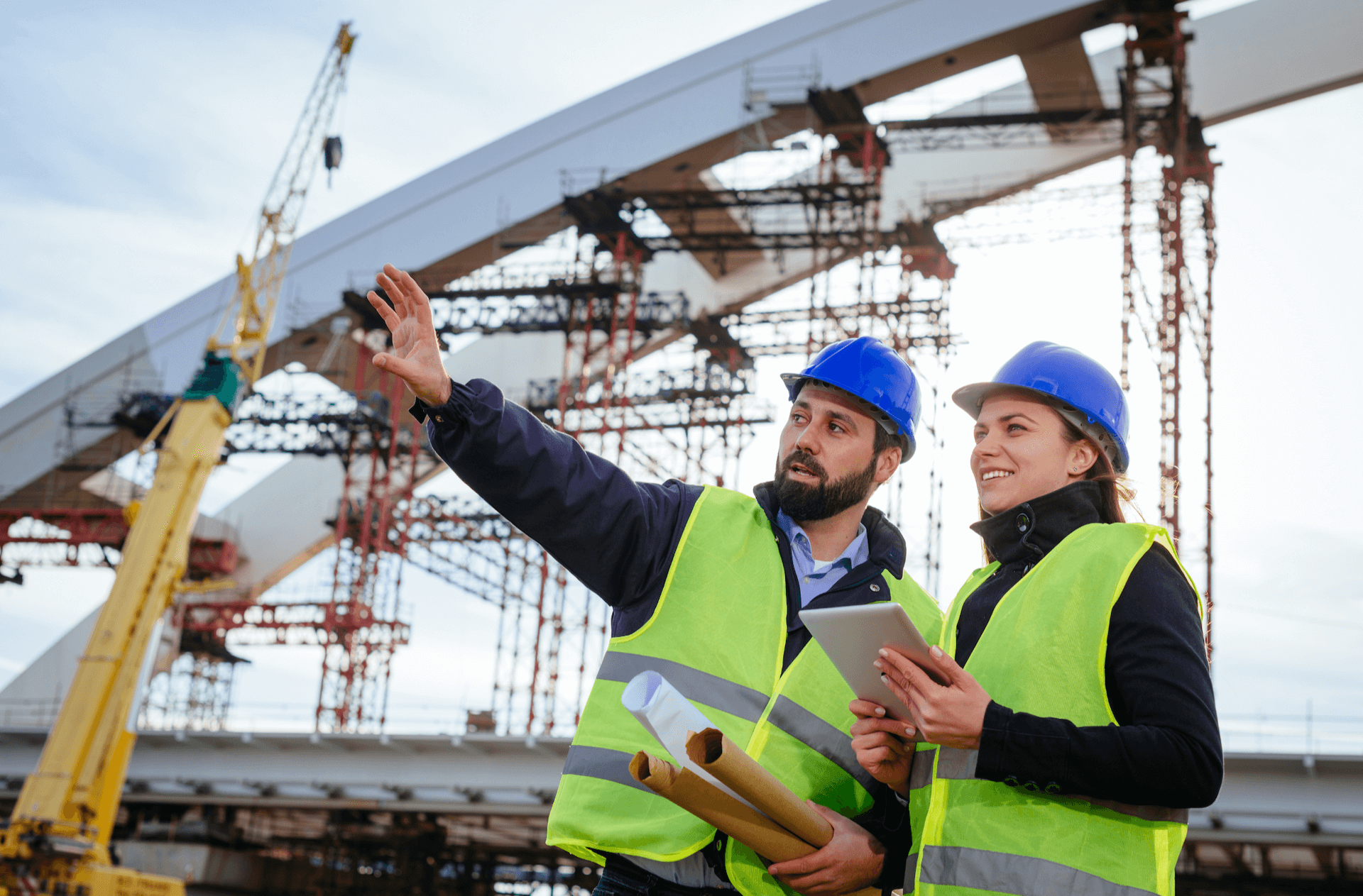 A man and women on a worksite outside. The man is pointing and explaining what he is gesturing towards to the woman.